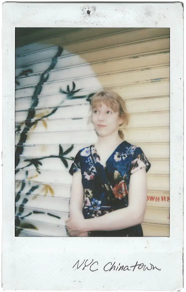 A Polaroid photo of Alexa, a young woman in a blue floral wrap dress. She leans against a garage door spray painted with an ink bamboo shoot. A handwritten caption reads 'NYC Chinatown'.